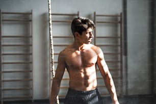 A fit young man in gym standing topless in front of a climbing rope. Copy space.