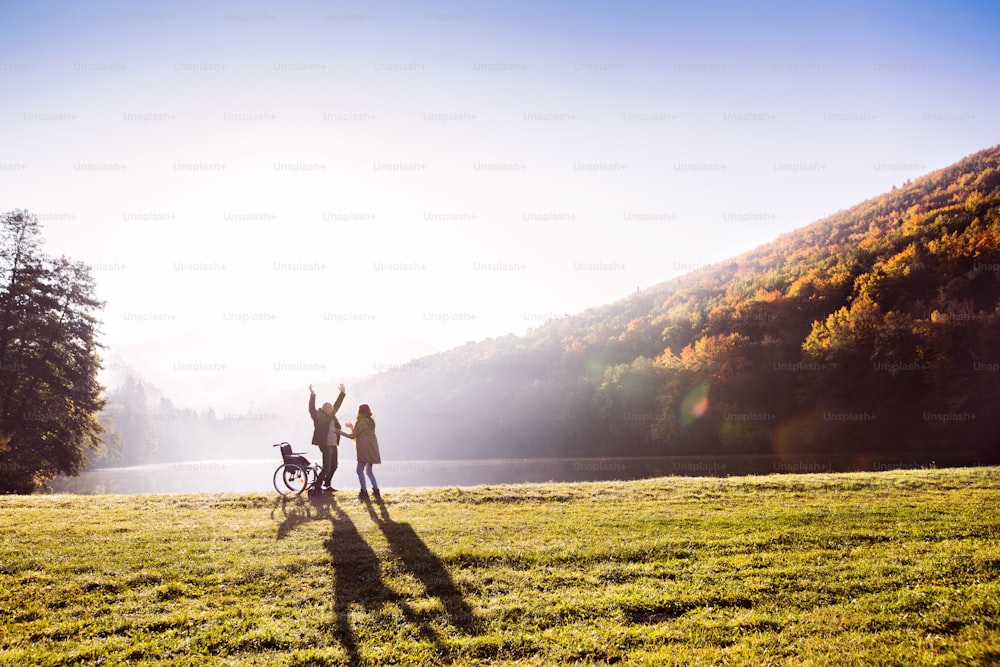 아름다운 가을 자연 속에서 호숫가를 산책하는 활동적인 노부부. 이른 아침에 휠체어 옆에 서 있는 여자와 남자.