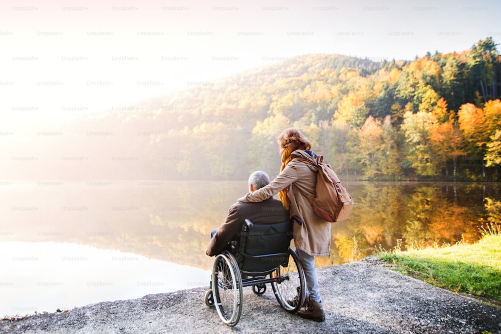 Aktives Seniorenpaar bei einem Spaziergang in einer wunderschönen Herbstnatur. Eine Frau und ein Mann im Rollstuhl am See am frühen Morgen.
