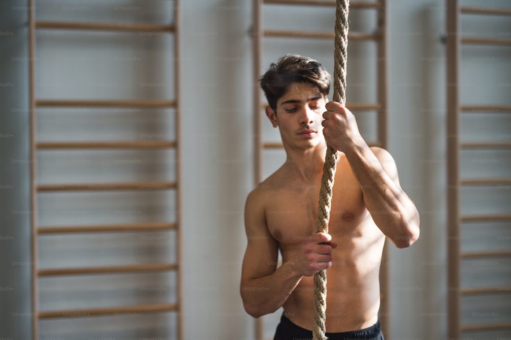 A fit young man in gym standing topless , holding a climbing rope. Copy space.