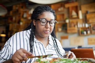 Hungry young woman in eyeglasses leaning over table with served pizza while slicing it