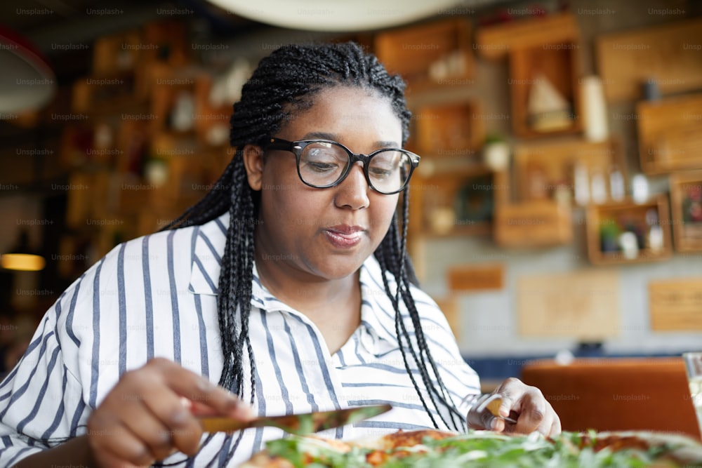 Hungry young woman in eyeglasses leaning over table with served pizza while slicing it