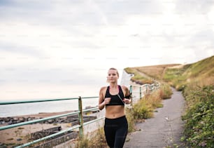 Young sporty woman runner with earphones running by the sea outside in nature, listening to music.