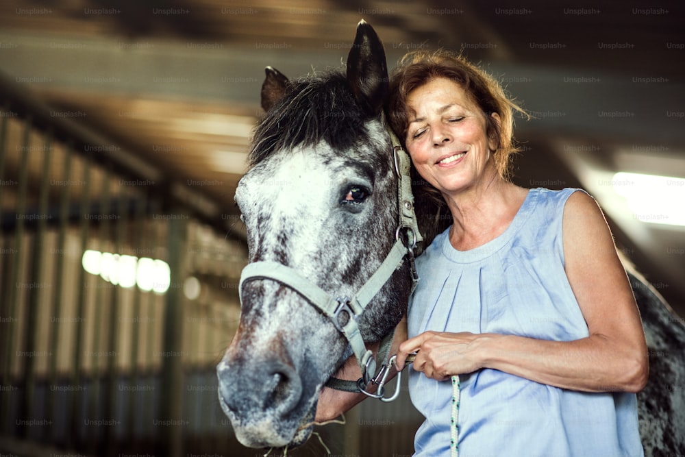 A happy senior woman with closed eyes standing close to a horse in a stable, holding it.