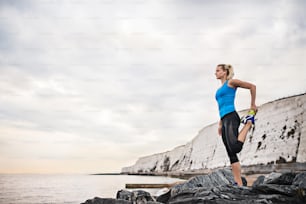 Young sporty woman runner with earphones stretching outside on the beach in nature. Copy space.