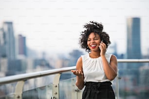 Un retrato de una mujer de negocios negra de pie contra el panorama de la vista panorámica de la azotea de Londres, haciendo una llamada telefónica. Espacio de copia.