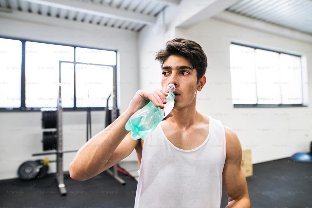 Junger, gutaussehender fitter Mann im Fitnessstudio, der sich zwischen den Übungen ausruht, Wasser trinkt.