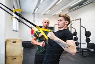 Fit young man with a personal trainer in gym working out, doing exercise with TRX.