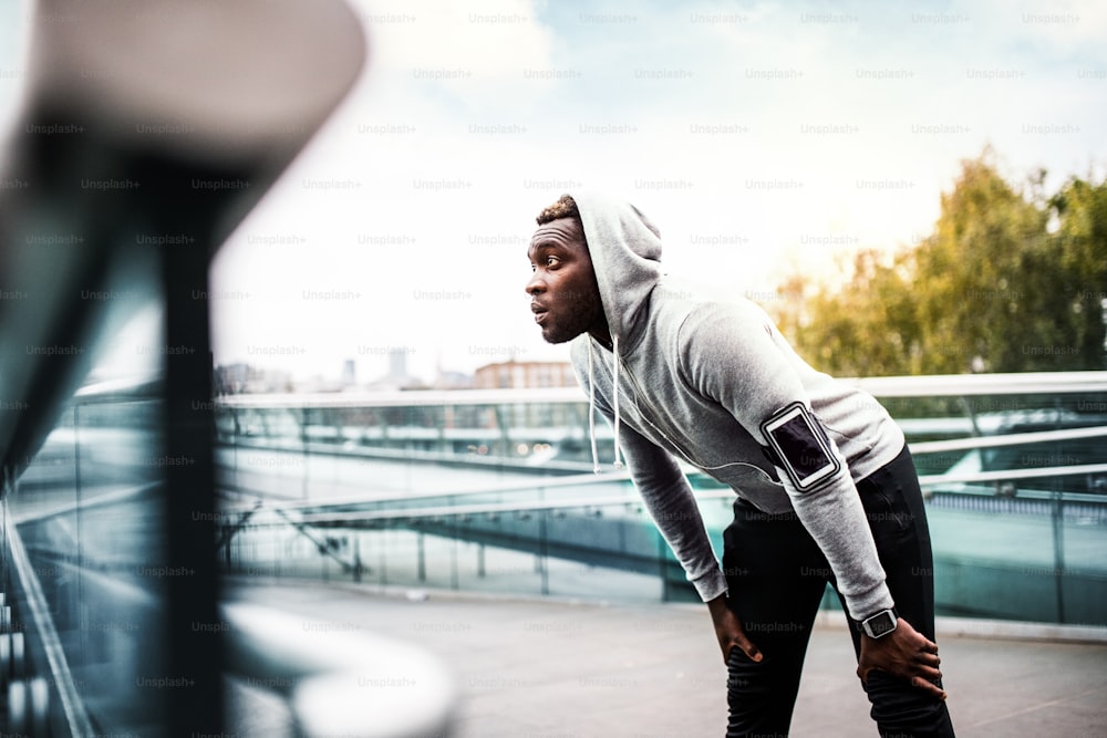Junger sportlicher schwarzer Mann Läufer mit Smartwatch, Smartphone und Kopfhörern auf der Brücke in einer Stadt, ruhend.