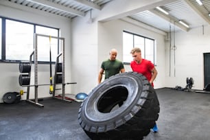 Fit young man with a personal trainer in gym working out, moving large tire.