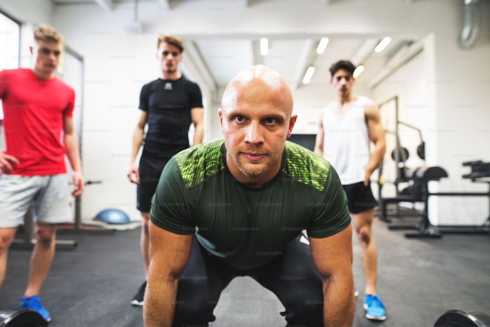 Three fit young men looking at a personal trainer in gym lifting barbell.