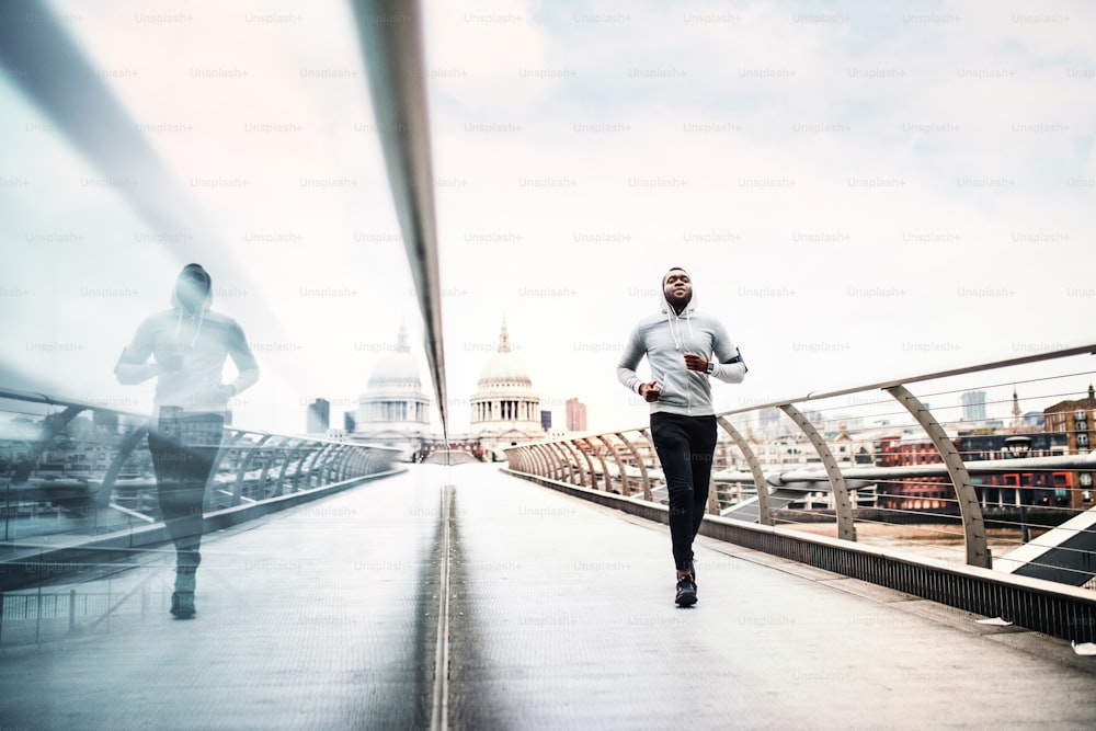 Junger sportlicher schwarzer Mann Läufer mit Smartwatch und Kopfhörern läuft auf der Brücke draußen in einer Stadt.