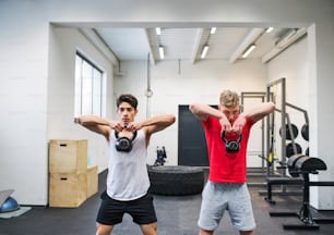 Two young fit men in gym gym working out, doing kettlebell swings.