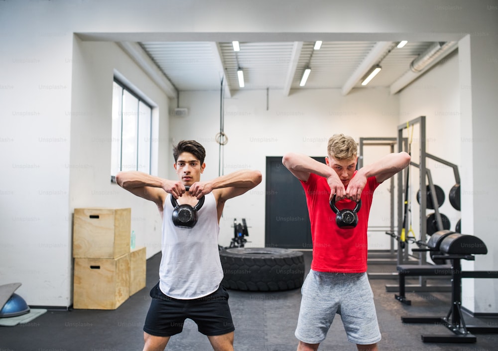 Two young fit men in gym gym working out, doing kettlebell swings.