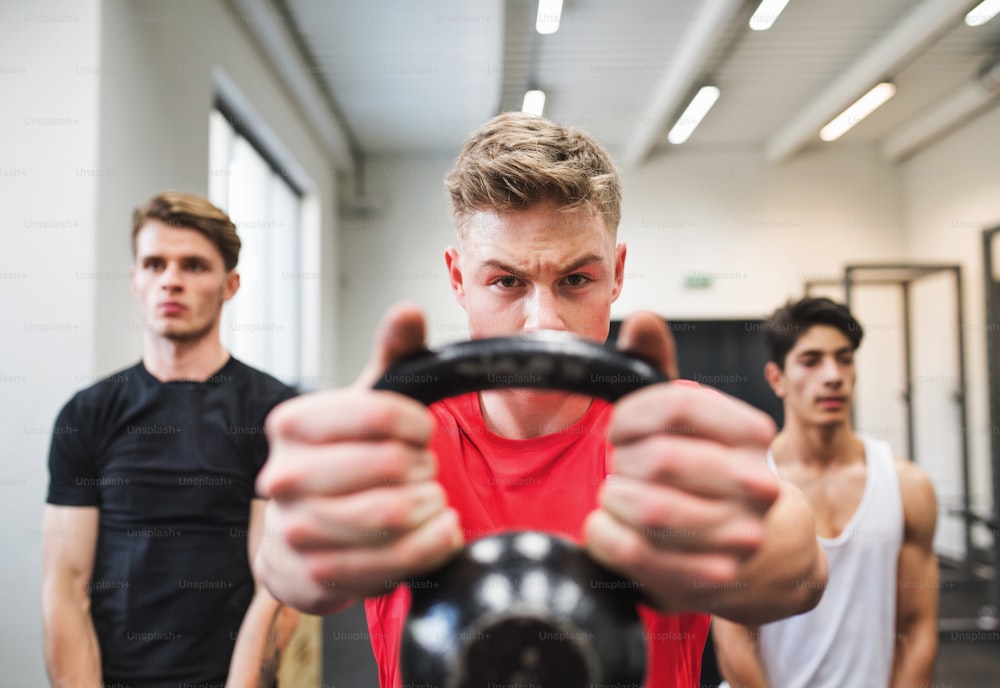 Three young fit men in gym gym working out, doing kettlebell swings.