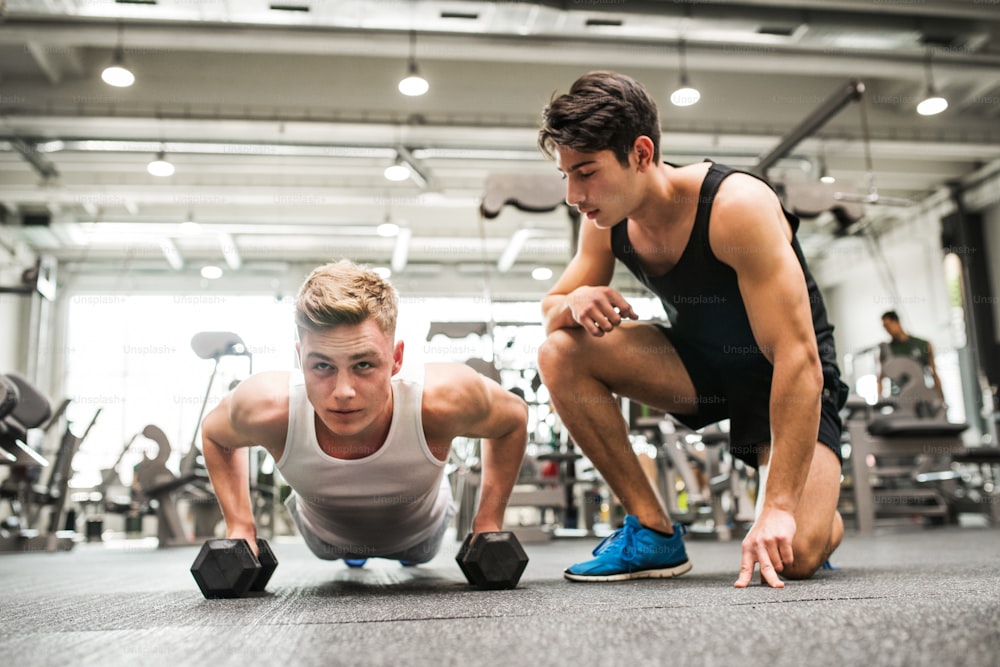 Young fit men friends in gym doing push ups on dumbbells, a friend supporting him.