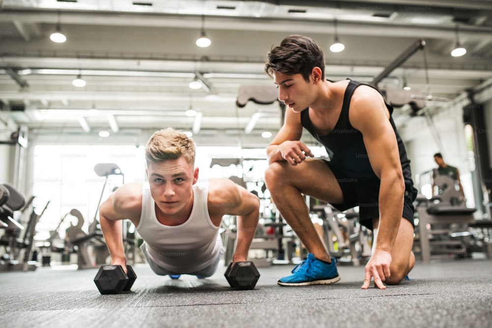 Junge fitte Männerfreunde im Fitnessstudio machen Liegestütze auf Hanteln, ein Freund unterstützt ihn.