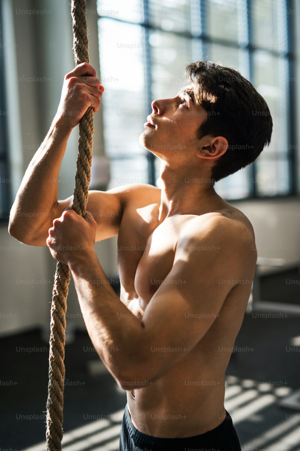 A fit young man in gym standing topless, holding a climbing rope.
