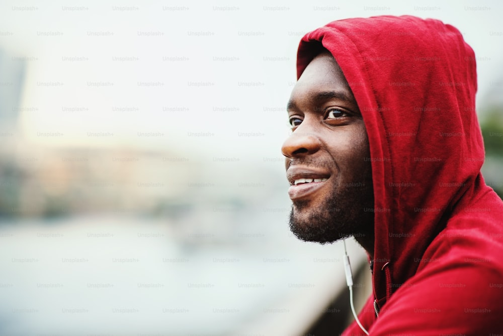 A close-up of black man runner with earphones and hood on his head standing in a city. Copy space.