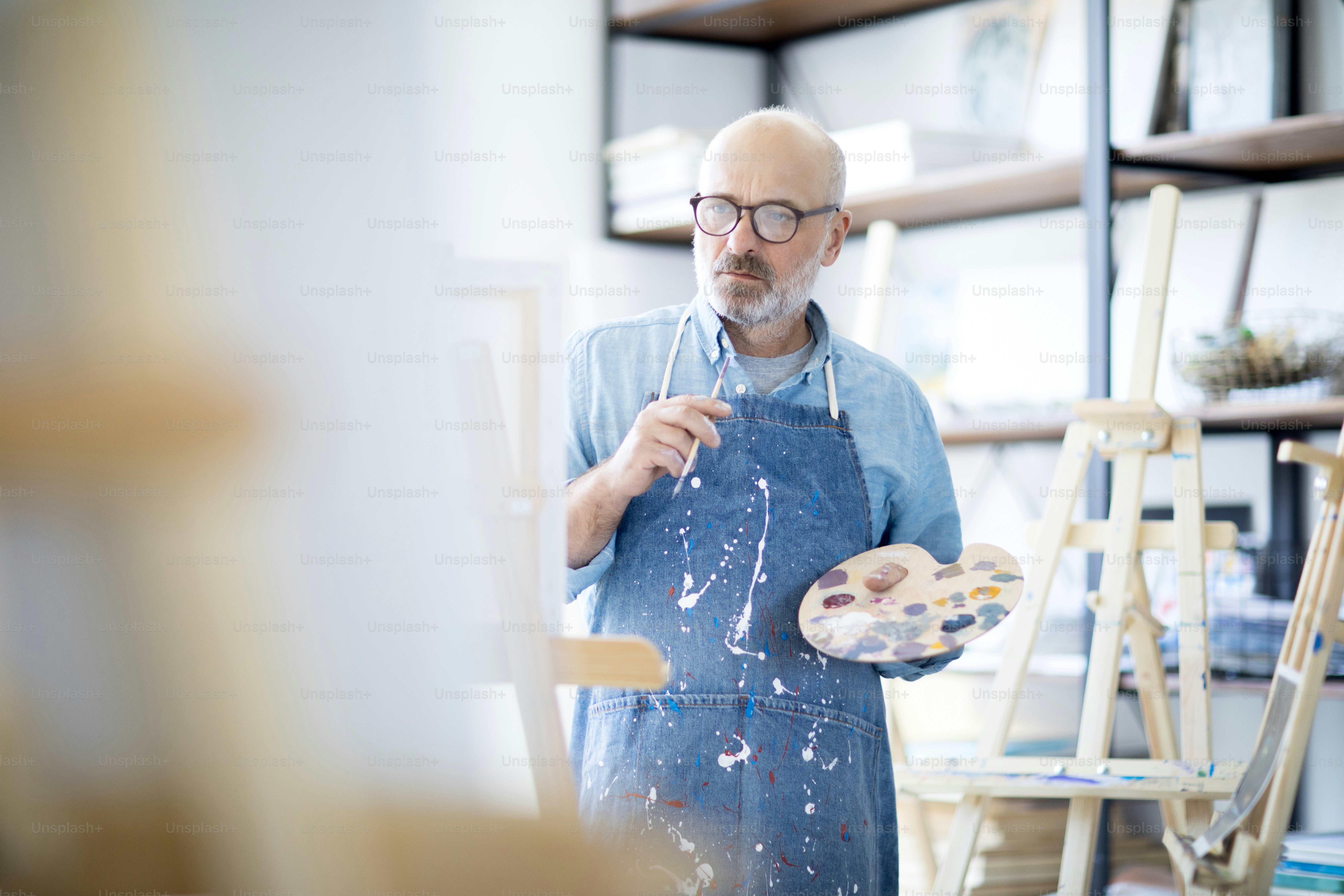 Serious and pensive mature man in apron looking at easel during creative process in studio