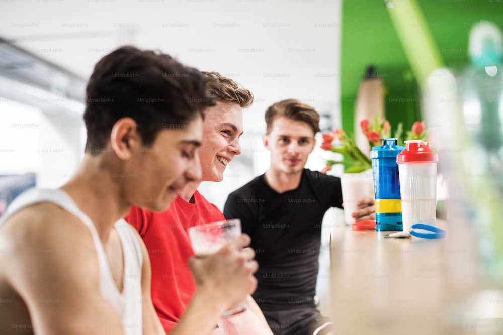 Three young fit men in gym gym sitting and holding glasses with protein drinks.