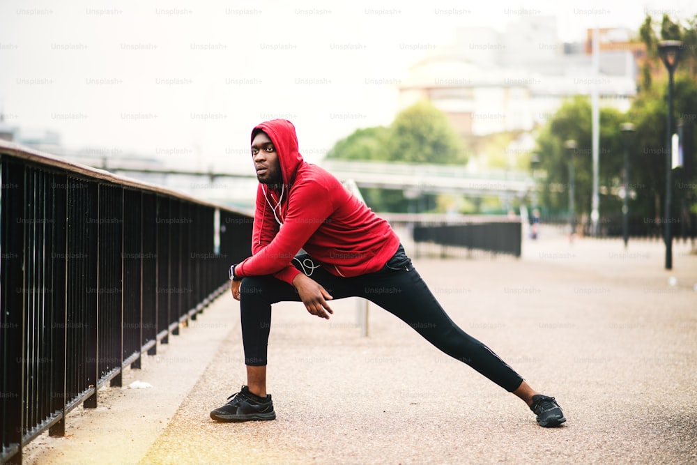 Young sporty black man runner with smartwatch and earphones doing stretching on the bridge outside in a city.