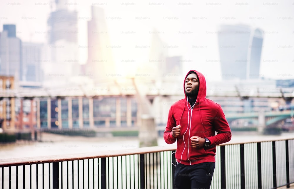 Young sporty black man runner with smartwatch and earphones running on the bridge outside in a city.