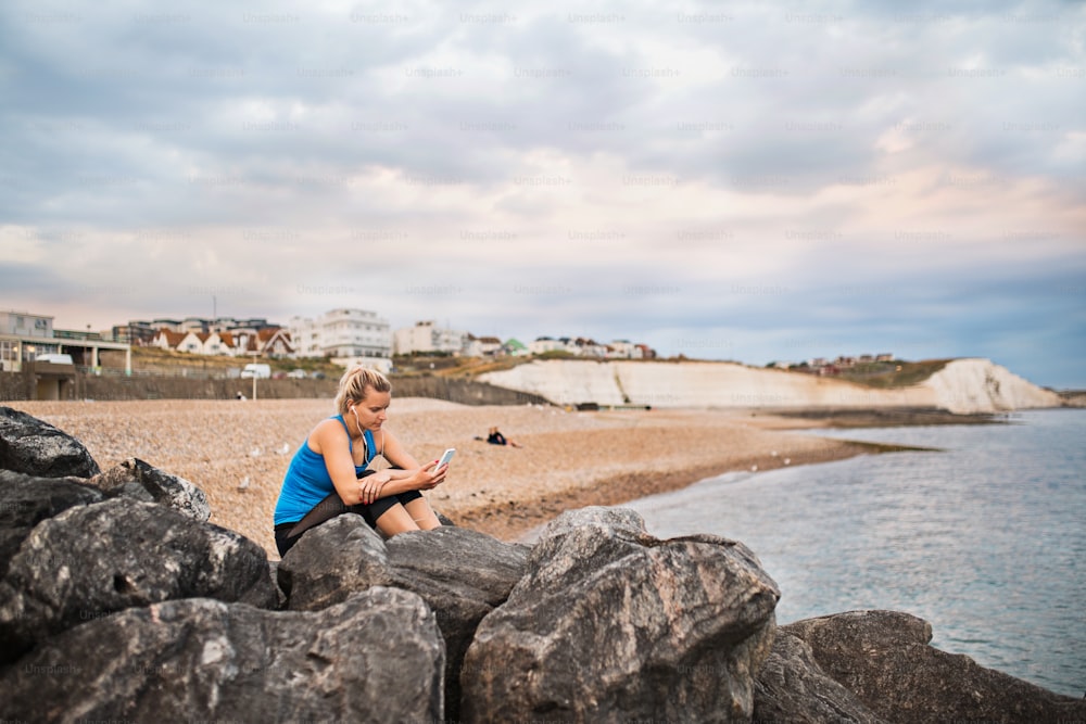 Young sporty woman runner with earphones and smartphone sitting outside on the beach in nature, listening to music and resting.