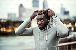 Young sporty black runner with smartwatch, earphones and smartphone in an armband on the bridge in a city, resting. A man putting a hood on head.