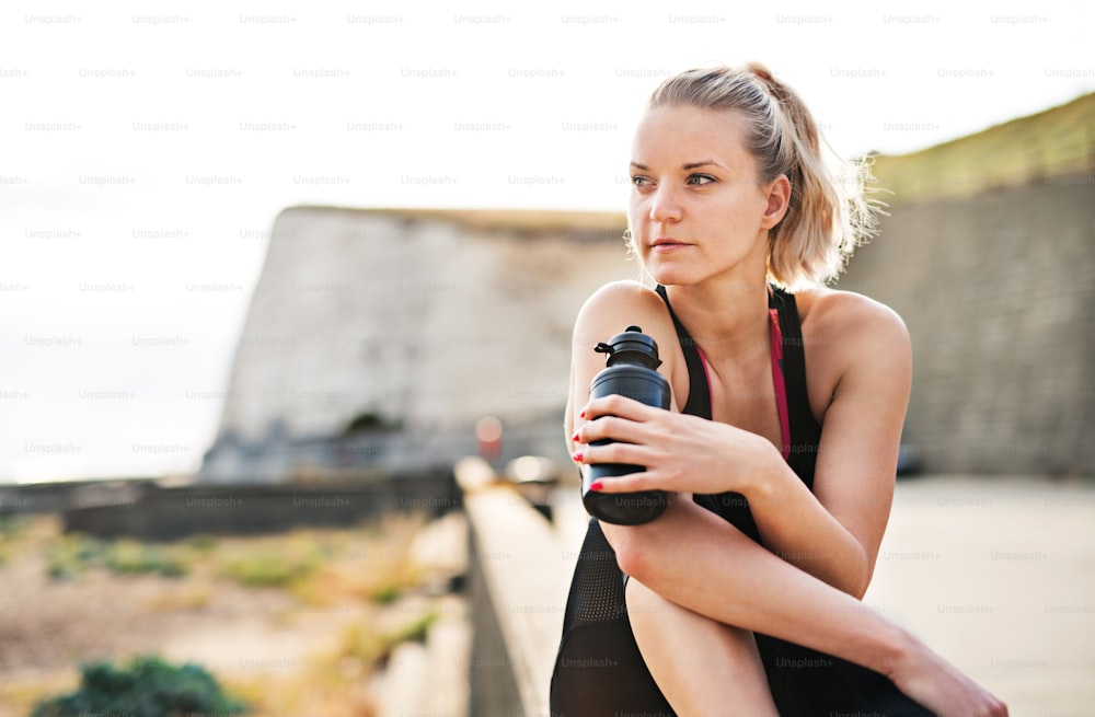 A close-up of young sporty woman runner with water bottle sitting outside, resting. Copy space.