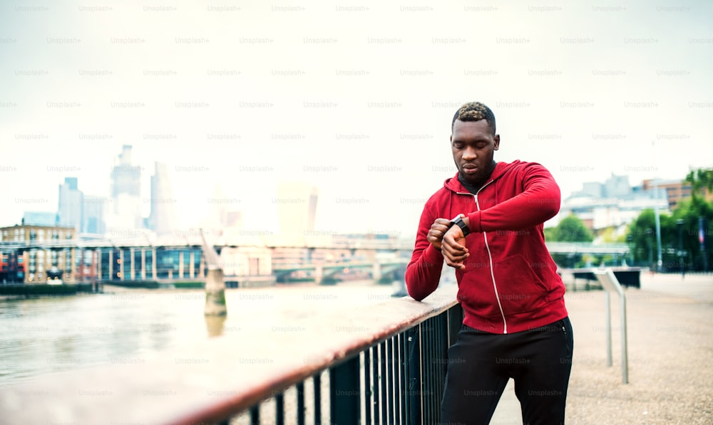 Young sporty black man runner setting smartwatch on the bridge outside in a city.