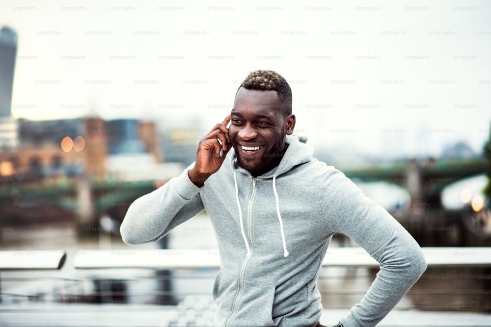 Junger sportlicher schwarzer Läufer mit Smartphone steht auf der Brücke in einer Stadt und telefoniert.