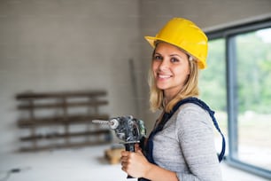 Portrait of a female worker holding an electirc drill on the construction site. Close-up of a beautiful young woman with a yellow helmet. Copy space.
