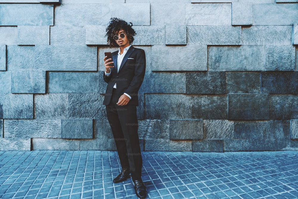 A stylish Asian guy in a formal suit with curly afro hair is standing in front of a textured wall and holding the smartphone; young Japanese businessman with the cell phone near the wall of blacks
