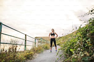 Young sporty woman runner in black activewear standing outside in nature, resting. Copy space.