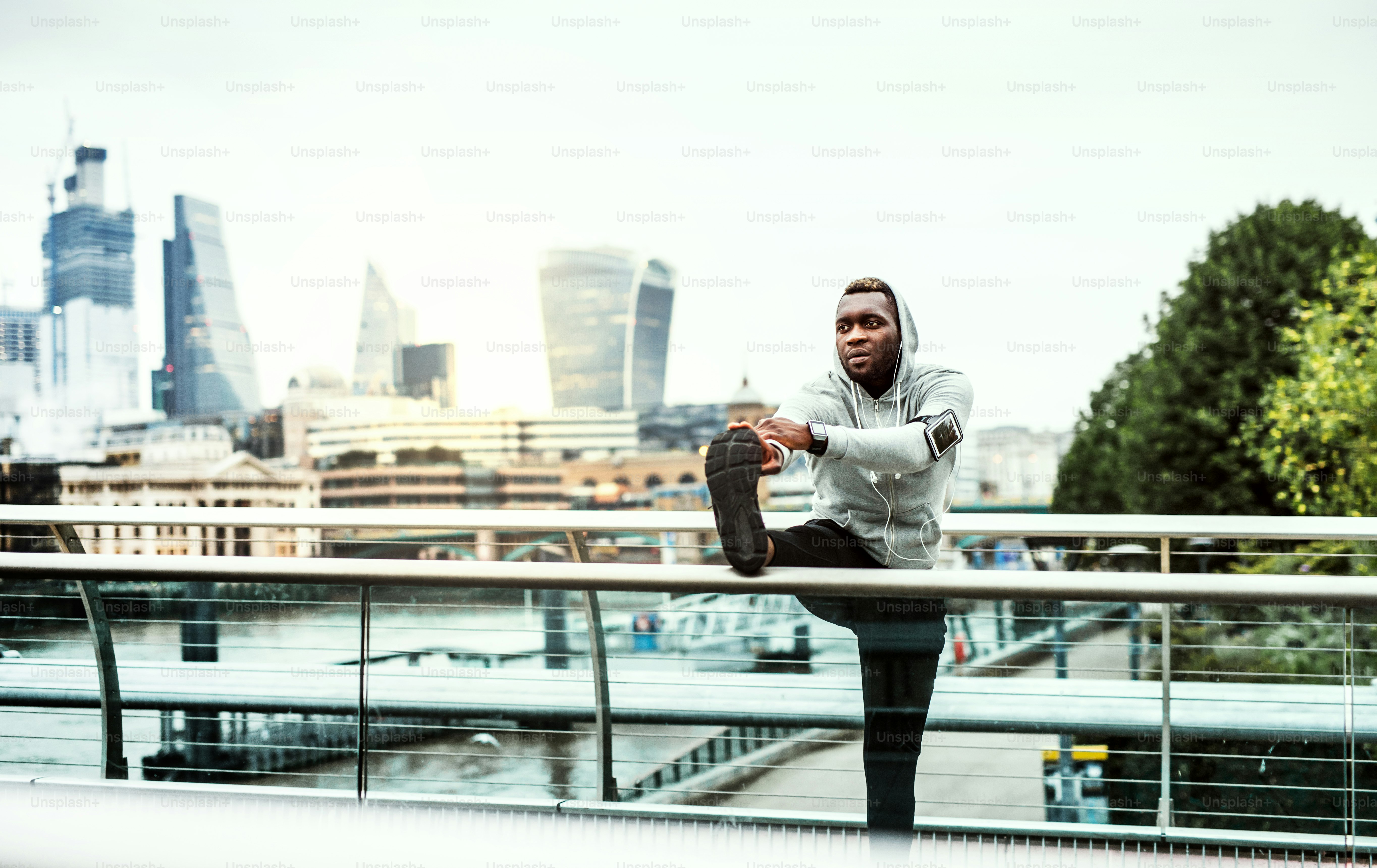 Young sporty black man runner with smartwatch, earphones and smartphone in an armband on the bridge in a city, stretching.