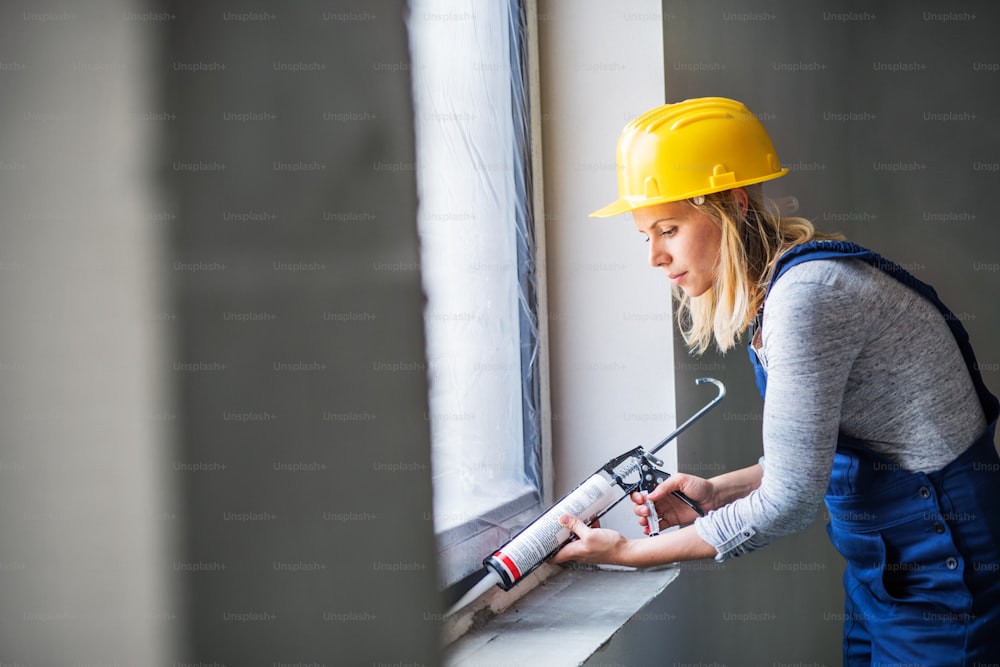 Young woman worker using silicone sealant gun on windows on the construction site.
