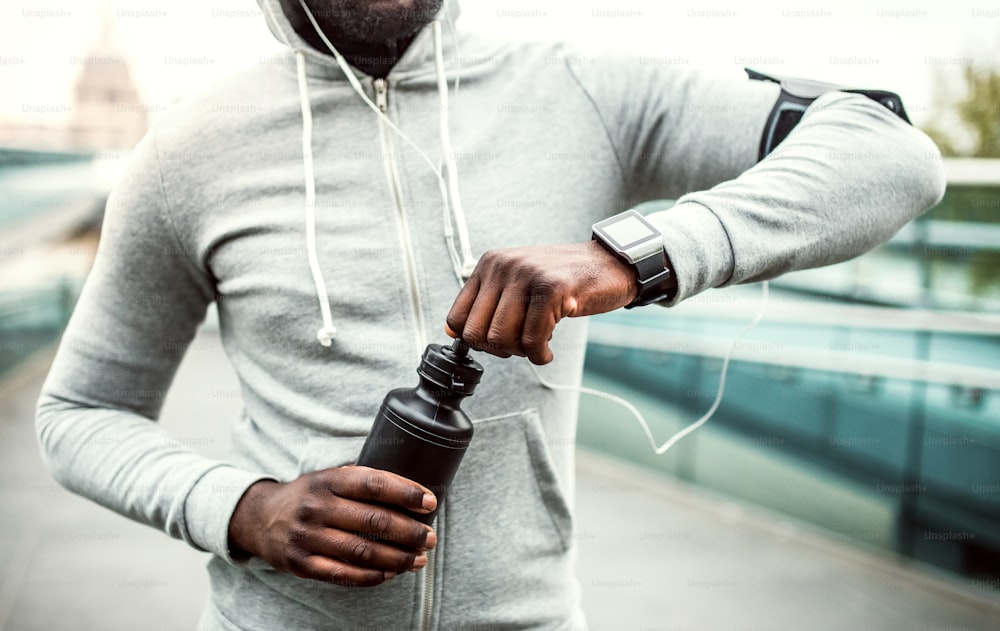 A close-up of unrecognizable young sporty black man runner with water bottle in a city.