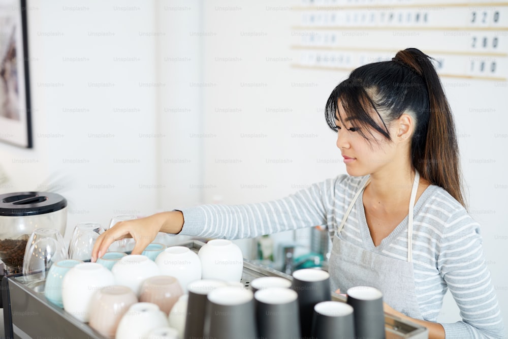 Mujer asiática joven en ropa de trabajo que pone tazas limpias en la bandeja mientras está de pie junto a la máquina de café