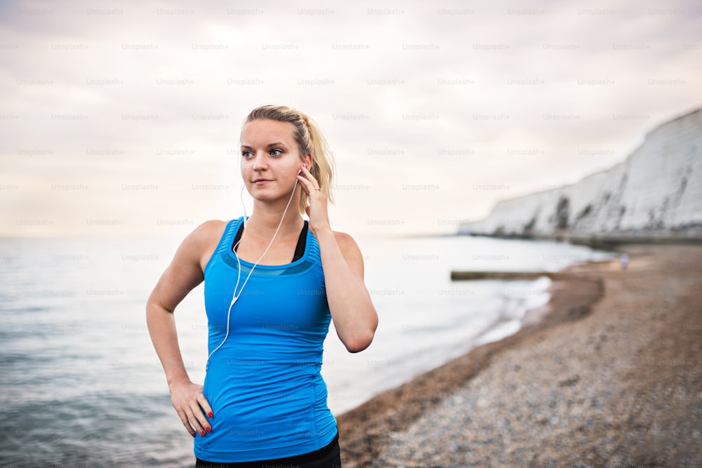 Young sporty woman runner with earphones standing outside on the beach in nature, listening to music and resting. Copy space.