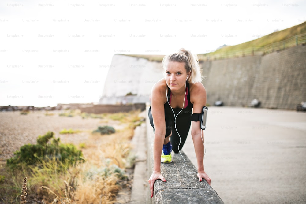 Jovem atleta mulher corredora com fones de ouvido e smartphone em uma braçadeira fora na natureza, alongamento.