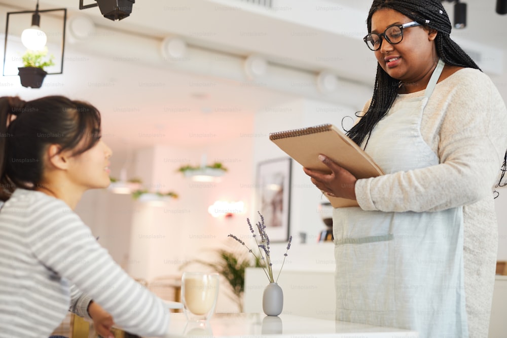 Young contemporary woman in workwear writing down order of client in notepad while standing in front of her