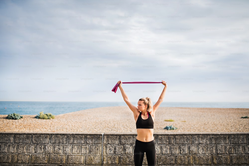 Giovane corridore donna sportiva che fa esercizio con elastici all'aperto su una spiaggia in natura.