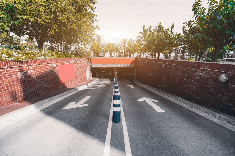 Wide-angle view of the outdoor entrance to a modern underground parking: two arrows showing the directions of entry, brick walls, traffic bollards, barriers, plenty of trees around; Barcelona, Spain