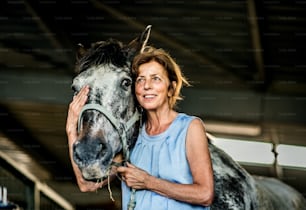 A happy senior woman with closed eyes standing close to a horse in a stable, holding it.