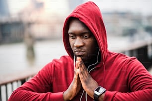 A close-up of black man runner with earphones and hood on his head standing in a city, stretching.