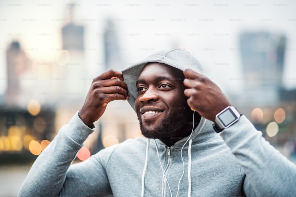 Young sporty black runner with smartwatch and earphones on the bridge in a London city, resting. A man putting a hood on head.