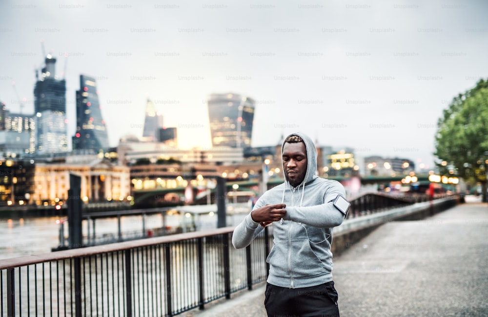 Young sporty black man runner with smartwatch, earphones and smartphone in an armband on the bridge in a London city, resting.