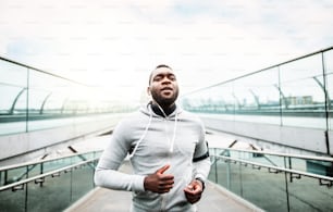 Young sporty black man runner with smartwatch and earphones running on the bridge outside in a city.