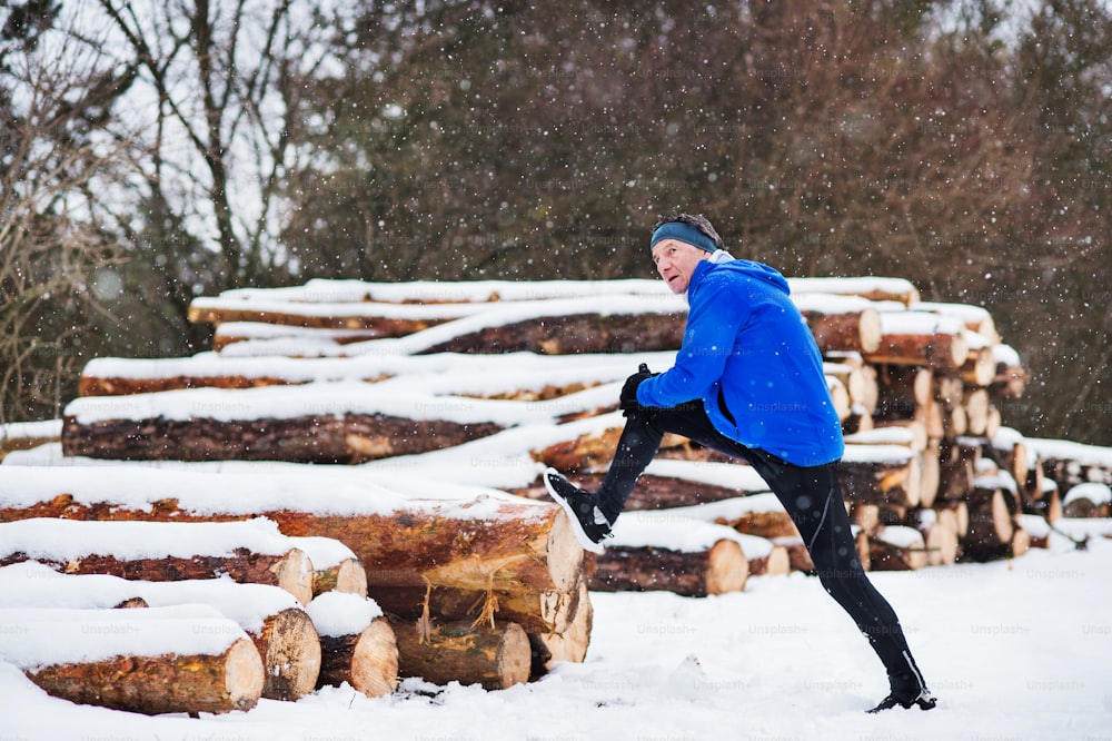 Ein älterer Mann, der die Beine vor dem Lauf in der winterlichen Natur am Baumstammstapel streckt. Speicherplatz kopieren.